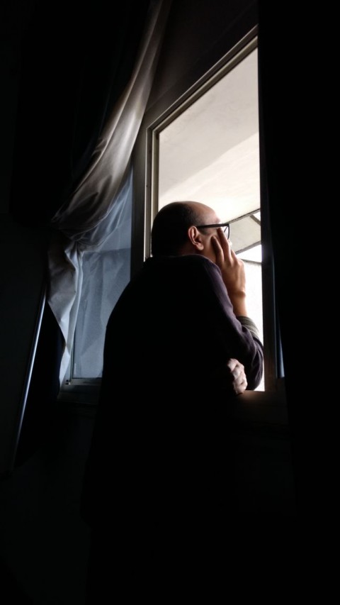 Photo of my uncle Aveek Sen, a middle-aged Bengali man, bald, with glasses, leaning over a windowsill and looking out to daylight, silhouetted, curtains billowing beside him, one hand under his chin, pensive.