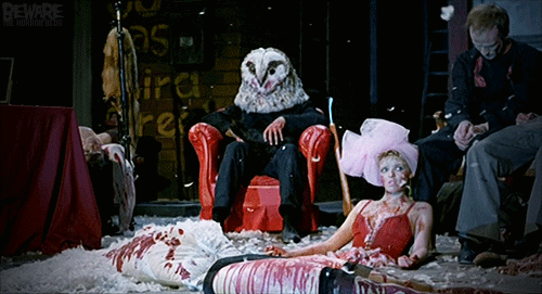 Gif from the film Stagefright, showing a spotlit man in black clothes and an owl mask sitting in a red chair on a stage, petting a cat, an axe propped by him, surrounded by bloody corpses arranged in a tableau, as feathers drift down like snow.
