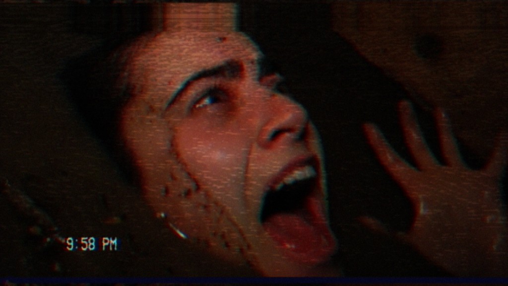 Shot from the film V/H/S/99, showing a young woman screaming in a closed, tight space, the footage grainy and blurred in the visual style of a camcorder from the 90s.