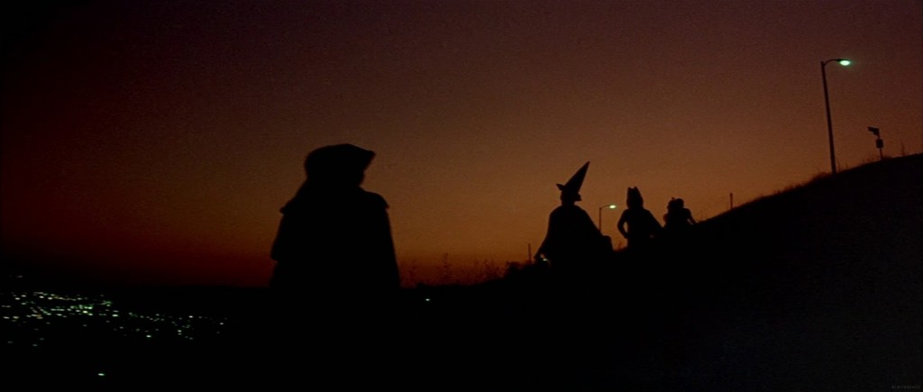 Shot from the film Halloween III, showing children in costumes silhouetted against an orange dusk sky and city lights.
